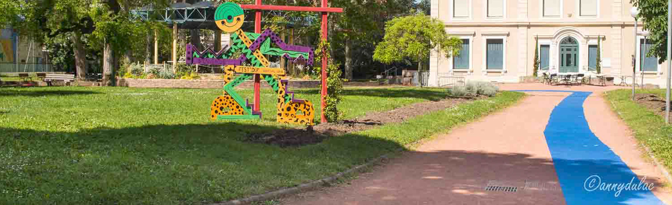 Image en plan large d'un parc citadin, couvert de pelouse, la statue du personnage représentant le festival Fest Dif est à gauche dans l'herbe. A droite, une allée de terre battue s'avance du bas de l'image jusqu'à un bâtiment au loin. Sur tout le parcours de l'allée est installé un cheminement de couleur bleue unie.